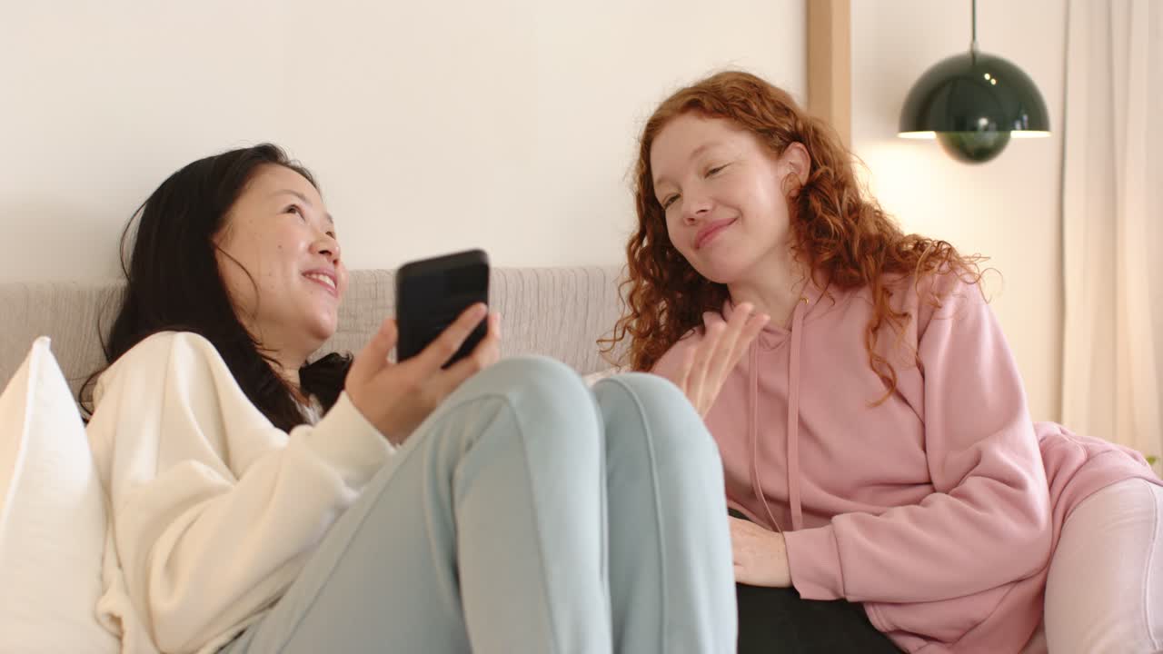Amigas diversas no sofá: a amiga à esquerda mexe no celular e mostra um clipe engraçado, a amiga à direita está rindo