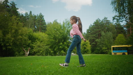 side view of young girl in pink shirt and jeans swinging pink balloon while walking on green grassy field with trees, parked yellow bus, and small vehicle passing by in background under blue sky