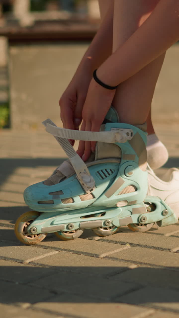 Lower view of individual sitting on bench wearing cyan roller skates while adjusting strap with black bangle on right hand, with white sneaker on pavement nearby, sunlight casts warm shadows