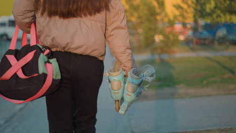 un retrato de una chica vestida con una chaqueta de melocotón y pantalones negros, llevando una bolsa de gimnasia y un par de patines mientras camina por un camino al atardecer