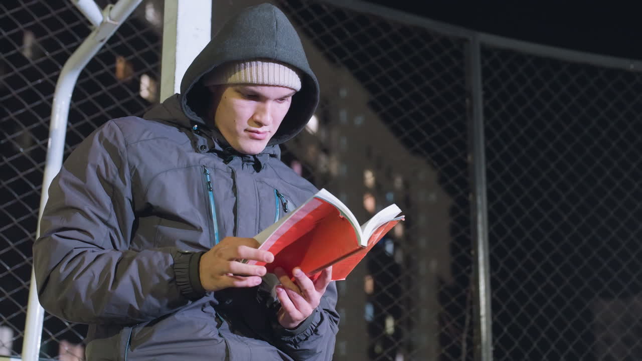 Man in hoodie and beanie leaning against metallic goalpost, contemplatively reading book outdoors at night, checking the pages under urban city lights with mesh fence background
