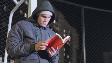 man in hoodie and beanie leaning against metallic goalpost, contemplatively reading book outdoors at night, checking the pages under urban city lights with mesh fence background