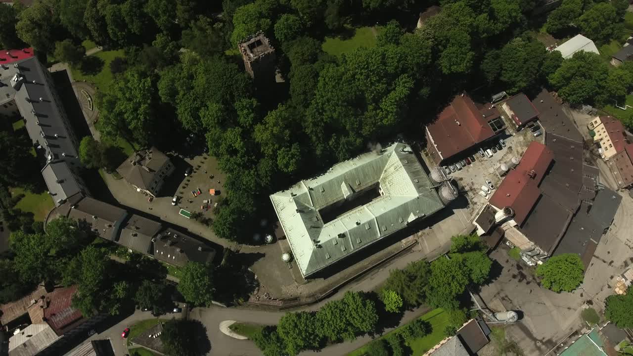 A building of an old brewery next to a park with a historic tower and a large number of green trees during summer in Poland, captured in a 4K bird's-eye view drone shot
