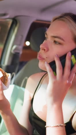 mujer de camiseta negra comiendo rosquilla mientras hace una llamada telefónica en un coche estacionado por la noche, mirando contemplativa, iluminación suave, bolsa de compras visible en el asiento trasero, con un ligero resplandor de los vehículos cercanos