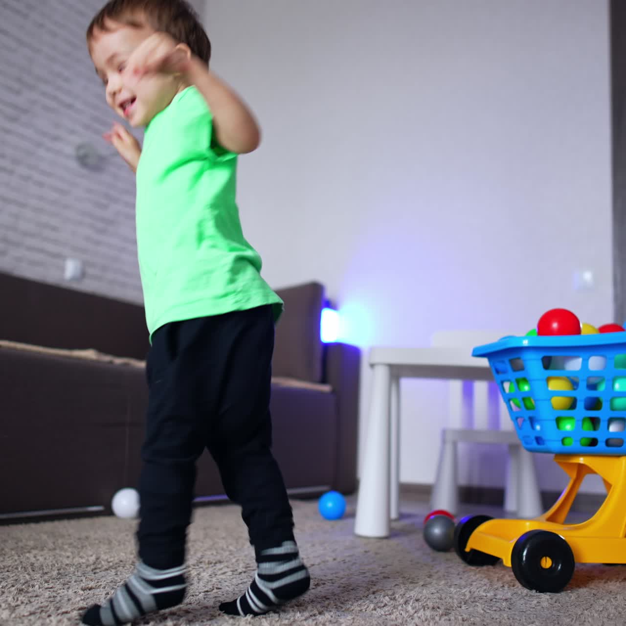 Happy energetic baby boy jumping and turning excitedly. Toy shopping cart and colorful balls near the kid in the room