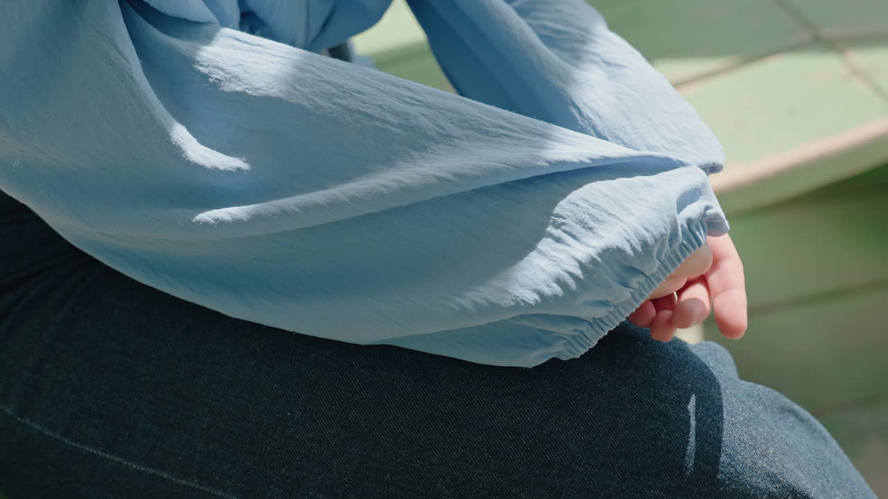 햇빛이 비치는 현관 벤치에서 파란색 셔츠 소매를 팔뚝 위로 걷어 올리고, 손으로 천을 다듬으며 활동을 준비하는 백인 여성의 모습이에요. 캐주얼한 데님과 은은한 그림자가 실용적이면서도 집중된 분위기를 연출합니다.
