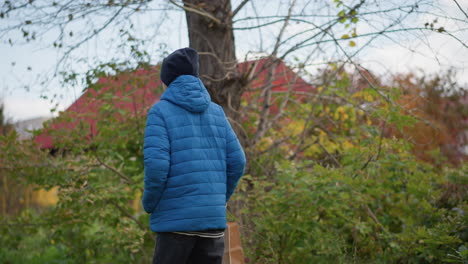 back view of person wearing blue jacket and black beanie walking outdoors with paper bag, surrounded by lush greenery and colorful autumn foliage, with distant view of red-roofed building