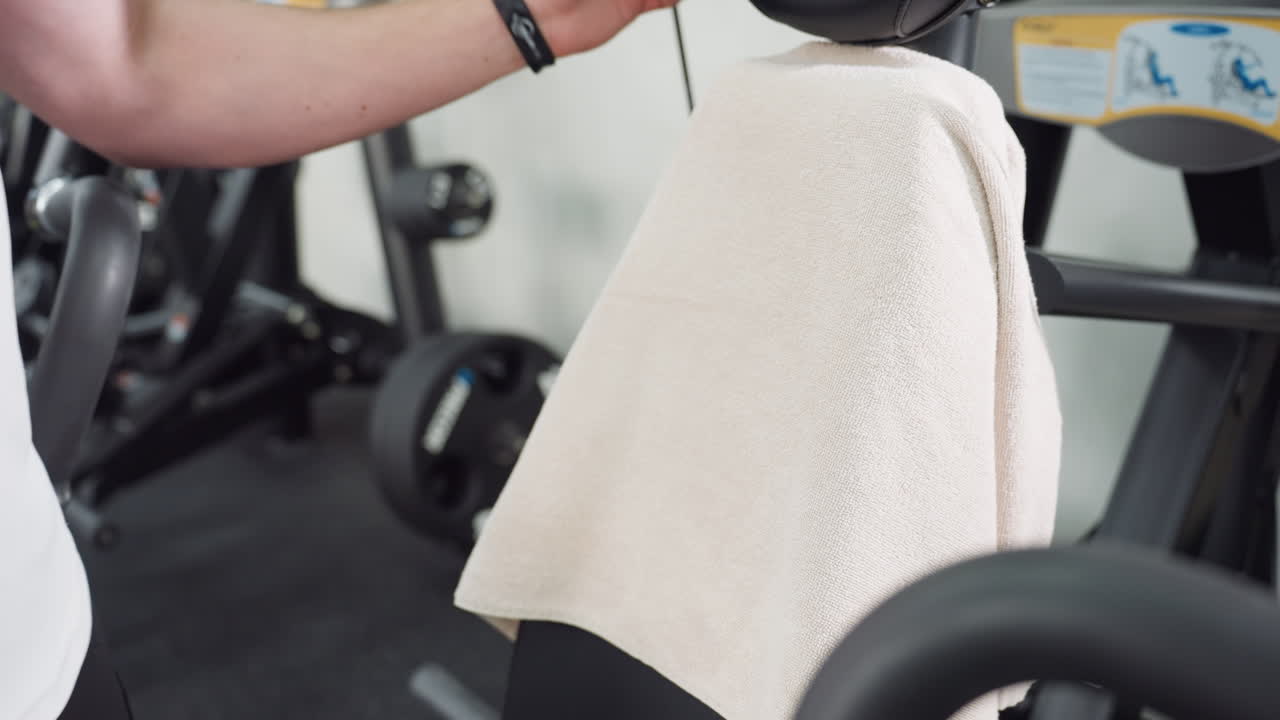 Young man drapes sports towel over rubber chair backrest before workout in modern gym with sleek weight machine and bright interior lighting preparing equipment for fitness routine session