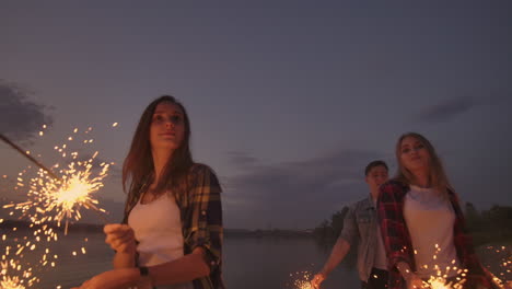 alegres amigos masculinos y femeninos están corriendo a lo largo de la playa al atardecer sosteniendo fuegos artificiales brillantes y luces fugitivas en cámara lenta. baile y fiesta de atardecer en la playa