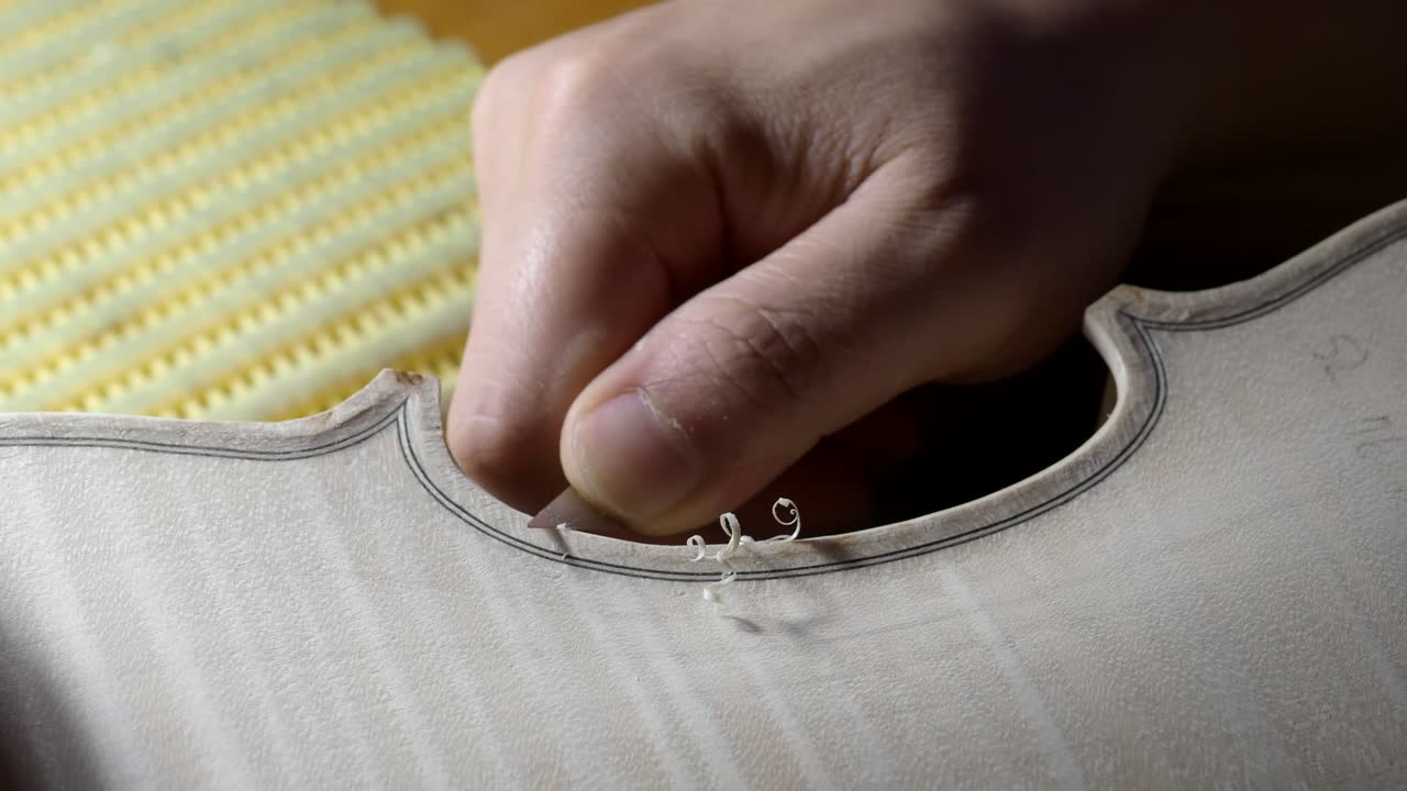 Extreme close-up shows luthier hand carving purfling channel and edges on violin top plate, with fine wood shavings curling away, capturing precision, tradition, and craftsmanship of violin making