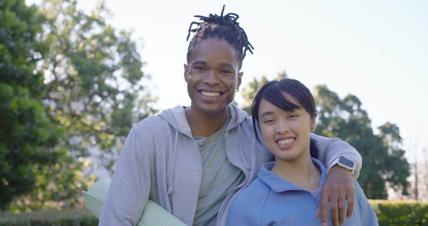 Et par, en afroamerikansk mand og en asiatisk kvinde, holder yogamåtter og er på vej til time, mens manden peger smilende.