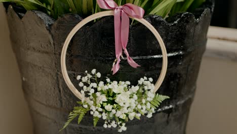delicado soporte de anillo de boda con flores blancas y cinta rosa, colocado contra una olla negra rústica