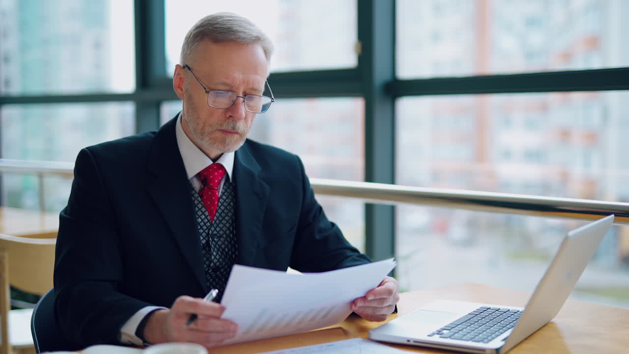 Portrait of senior businessman in formal costume. Mature man sitting at the table in front of laptop and working with papers in office near the window.