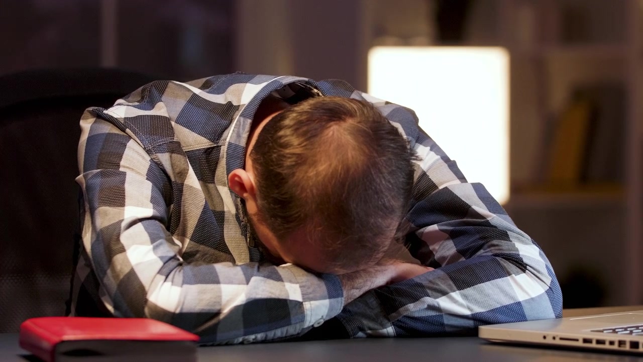 Hombre exhausto durmiendo en su escritorio en una oficina con poca luz