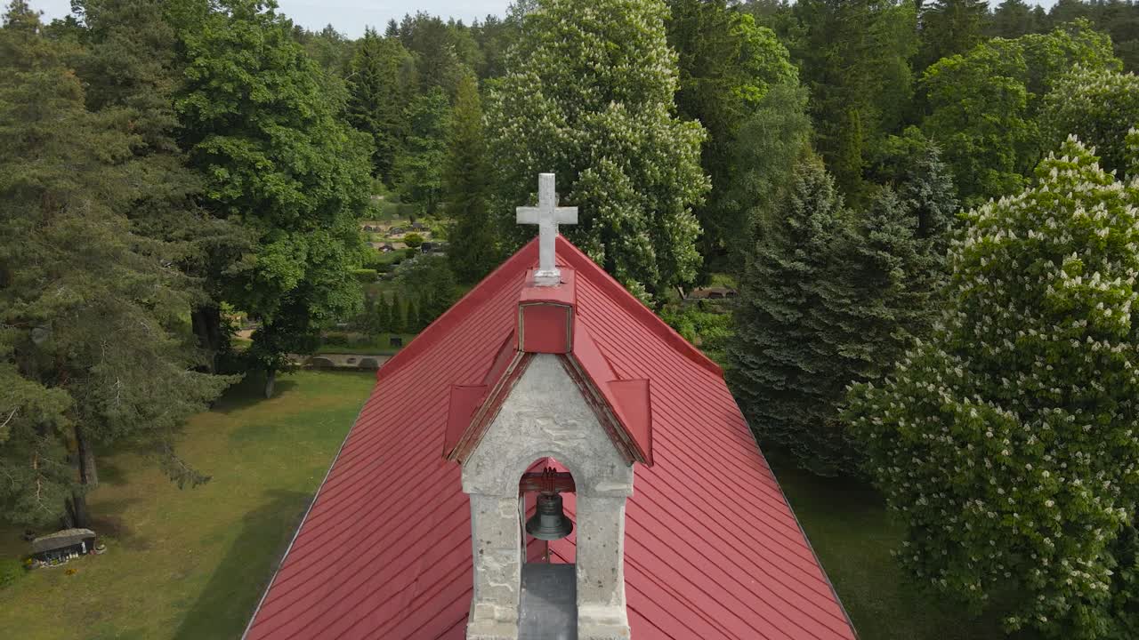 Aerial drone orbiting view of a rural white countryside church during a cloudy day in a forest. Religious building has a red roof and a white cross on top of it while horizon and treetops visible.