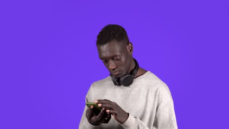 a young african american man wearing headphones on neck is typing on his smartphone standing isolated over blue wall background