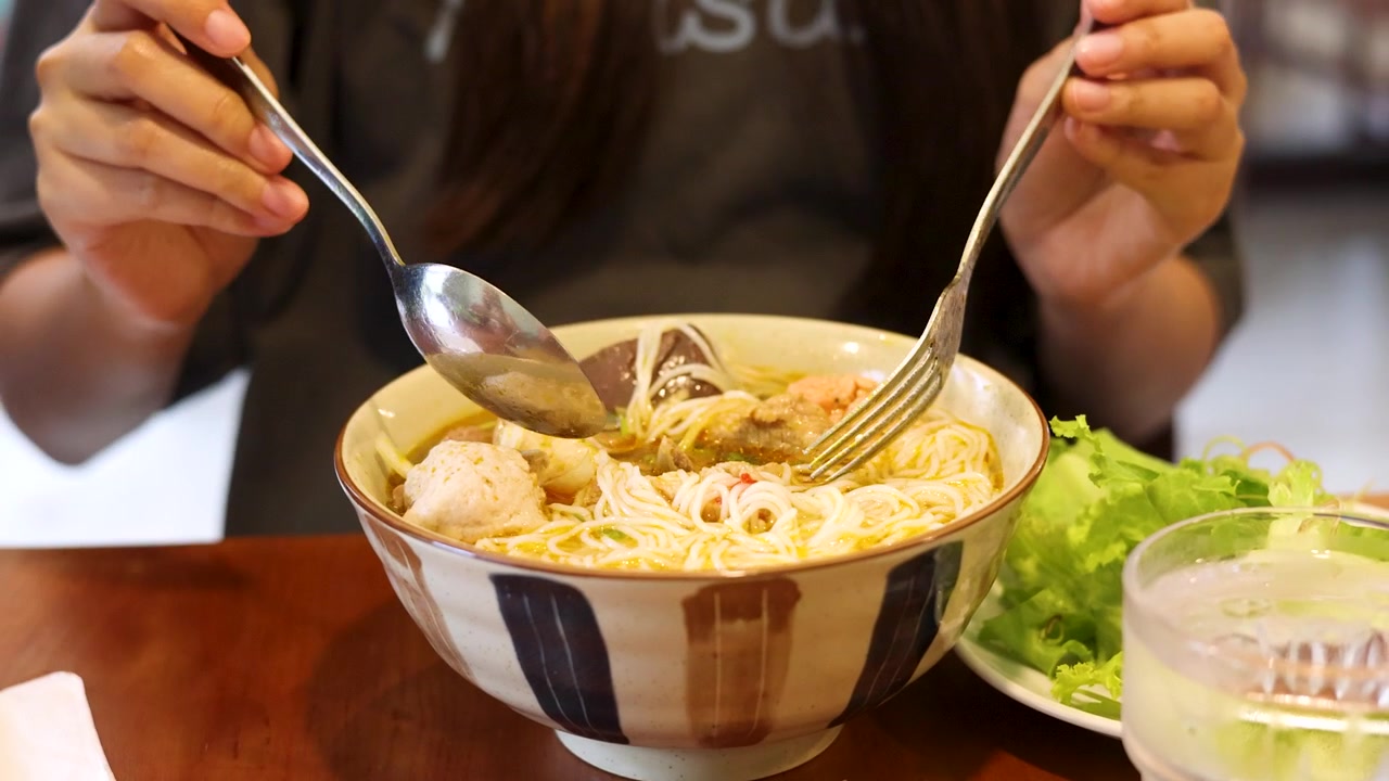 Close-up shot of a person using a spoon and fork to eat Vietnamese noodle soup