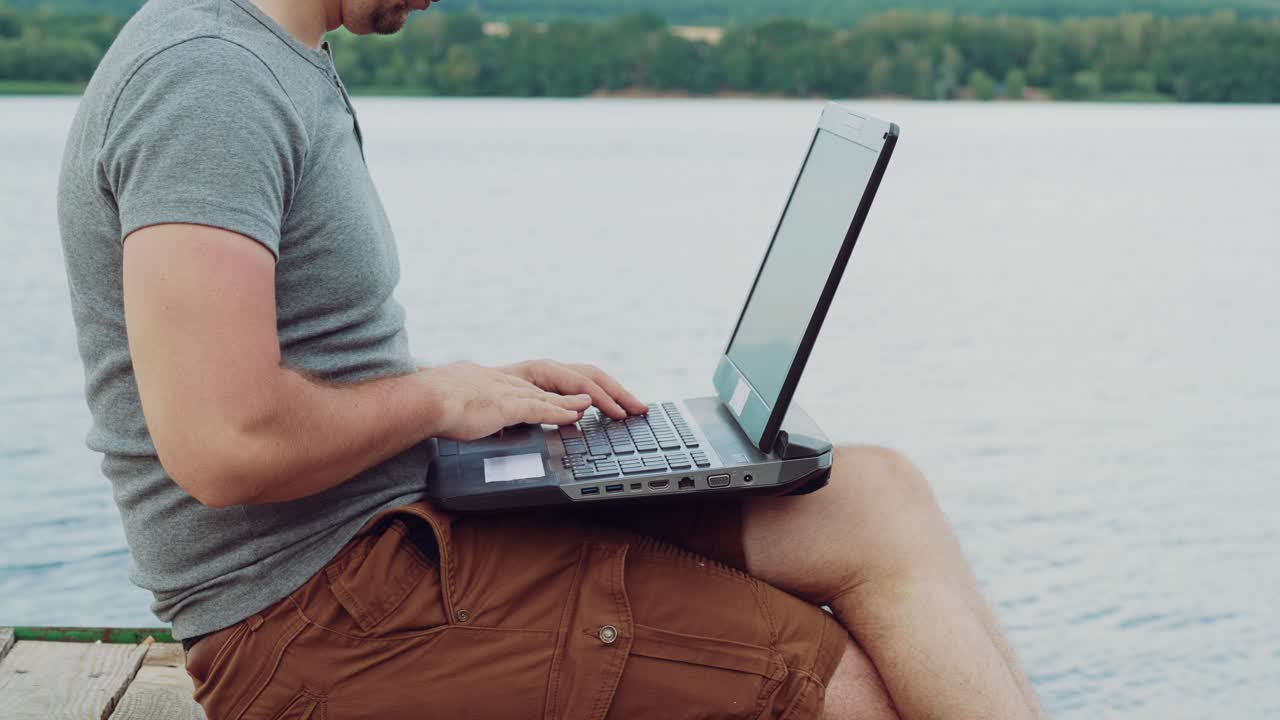 busy man in a gray t-shirt is sitting on a masonry and typing text behind a laptop on the background of nature