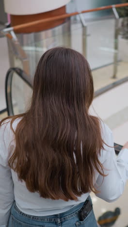 vista trasera de una joven adulta mostrando su cabello en la escalera mecánica descendente con la mano en el riel en un centro comercial ocupado, la gente moliendo alrededor en el fondo