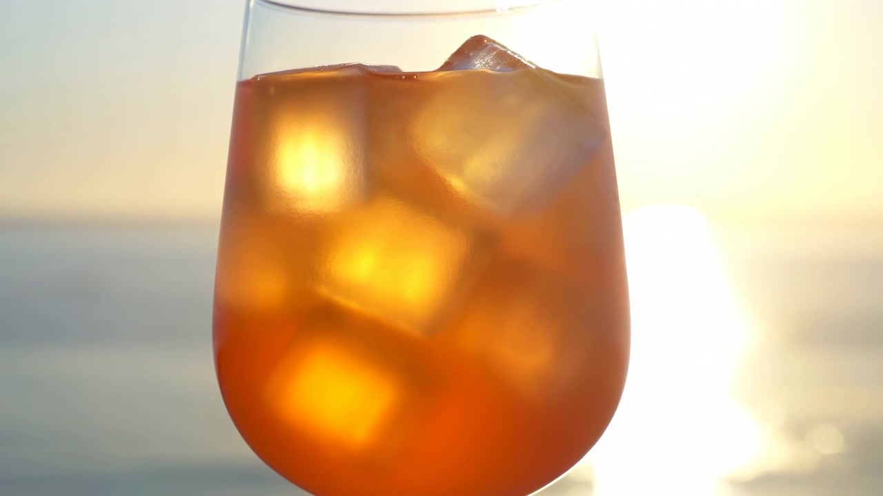 Grandi cubetti di ghiaccio brillano attraverso un liquido arancione mentre una calda retroilluminazione crea riflessi luminosi e punti luce traslucidi, con acqua blu calma e cielo pallido leggermente sfocati sullo sfondo.