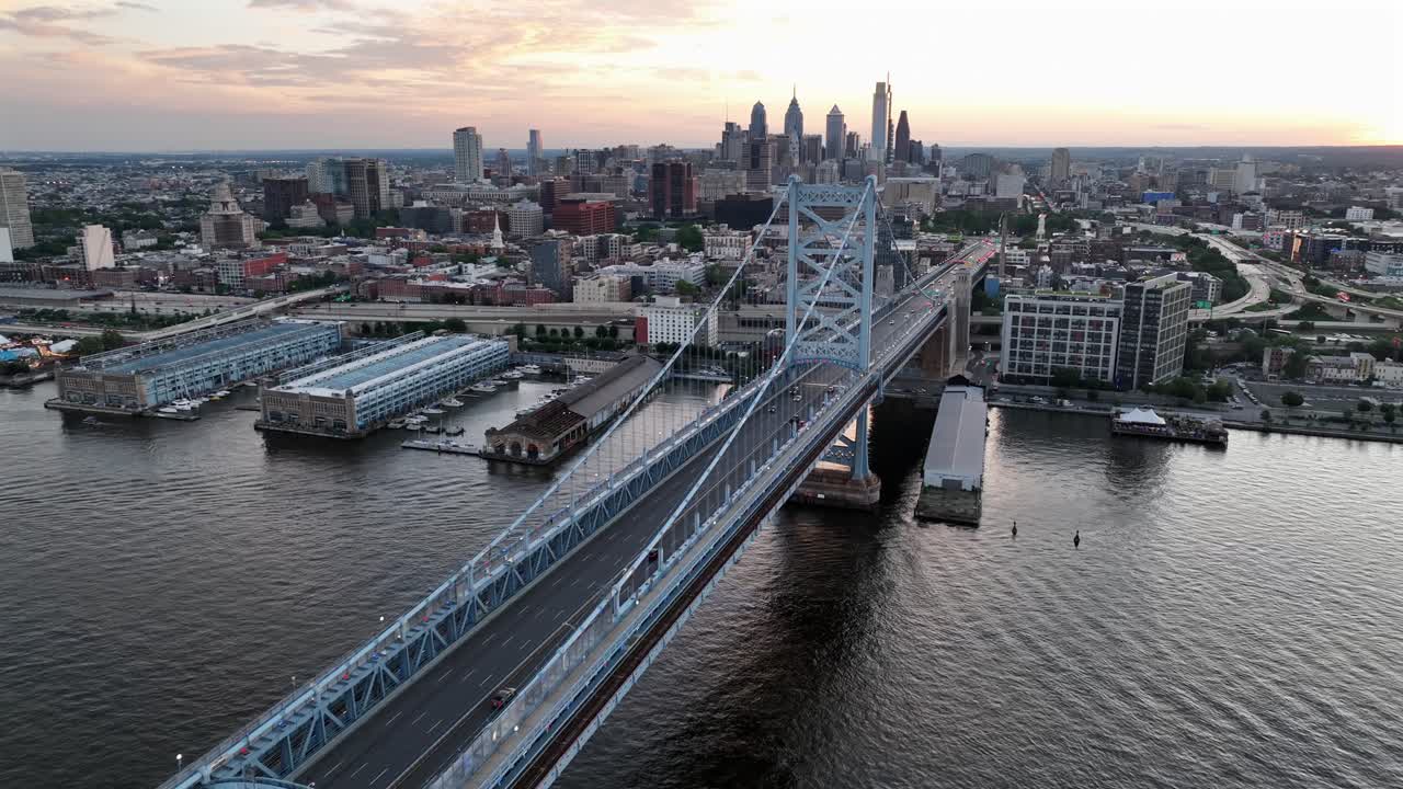 Cars on famous Benjamin Franklin bridge crossing river at dusk. Sunset time on Philadelphia. Skyline downtown with high-Rise buildings on background. Marina pier on waterfront