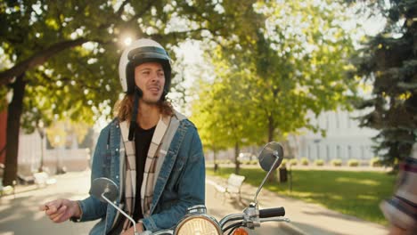 a brunette girl in a checkered shirt pats her boyfriend on the back and leaves him. a guy in a denim shirt with long and curly hair in a white helmet sits on a moped and makes a sad grimace and smokes a cigarette in the summer city