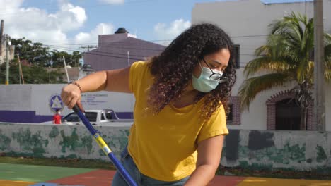 Young-Mexican-girl-with-abundant-curly-hair-wearing-glasses-and-masks-while-painting-the-ground-of-a-public-park-in-a-Mexican-neighborhood