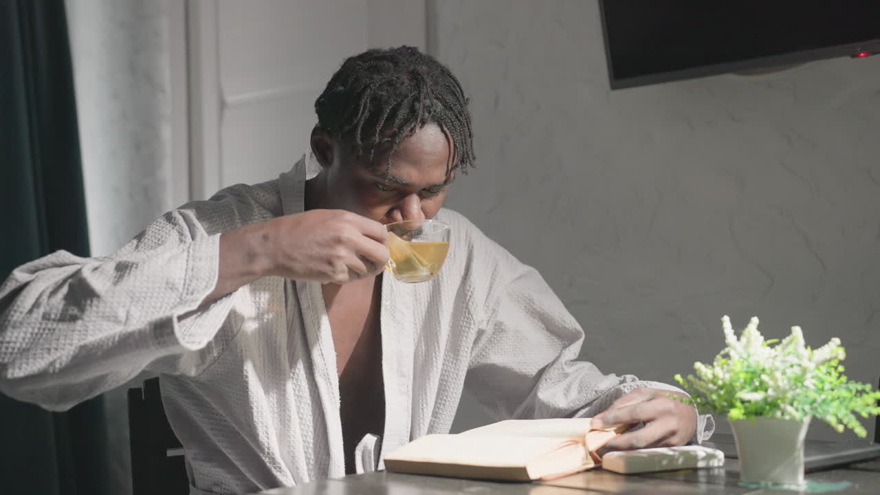 Man in white robe sitting at table with open book and tea, sipping from cup while focused on reading, relaxing moment in bright, cozy home setting