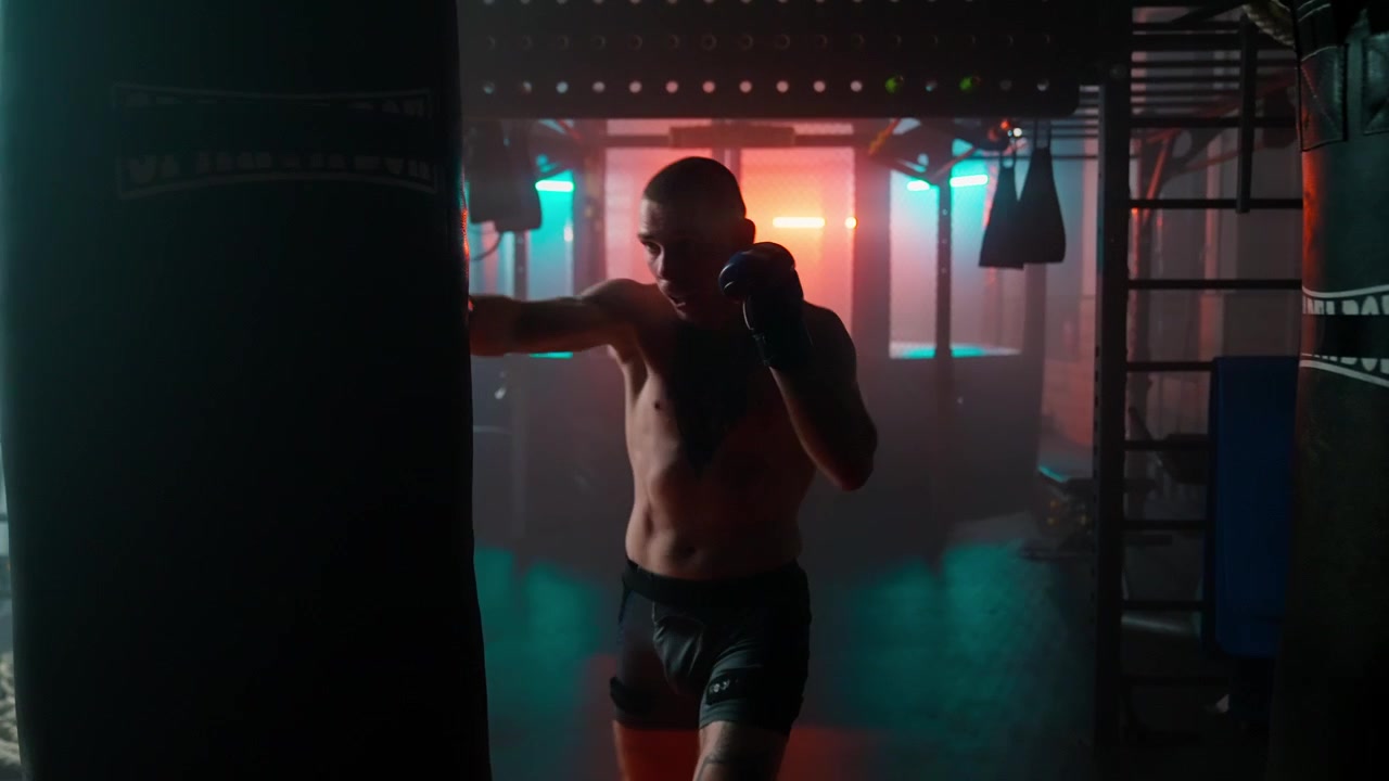 Shirtless Male Boxer Training with Punching Bag in Dark Gym with Neon Lights