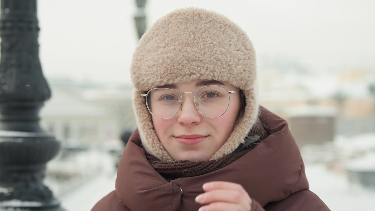 Ritratto centrato di giovane donna all'aperto in un parco urbano innevato, mani che si alzano per sistemare occhiali trasparenti mentre la neve si accumula su un soffice cappello beige, ciglia, spalle e cappotto sotto una luce soffusa e nuvolosa.