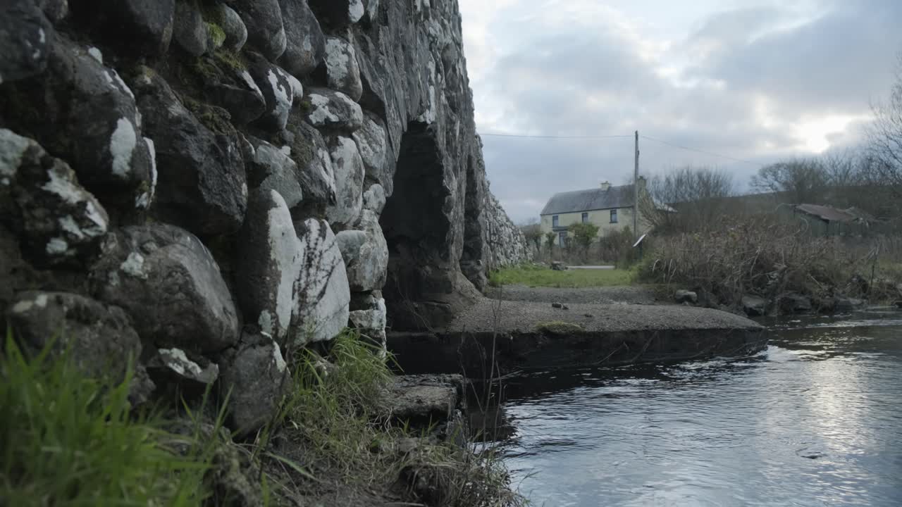 Close up shot of a river running under a stone bridge in Connemara, Ireland, Rack focus.  Small house in the background. Camera dollies backwards. Sun breaking through the clouds.
