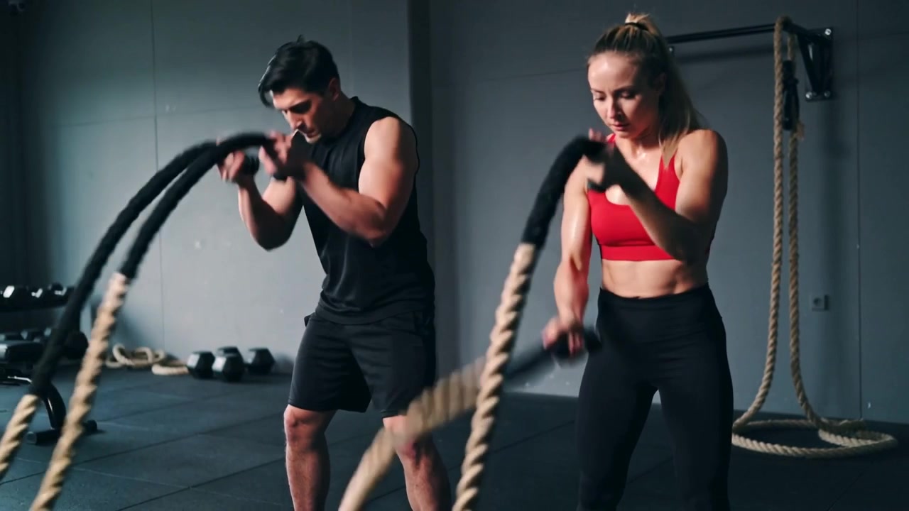Dos deportistas con equipamiento negro y rojo, fortaleciendo el core y ejercitándose con cuerdas de batalla en un gimnasio para desarrollar fuerza.