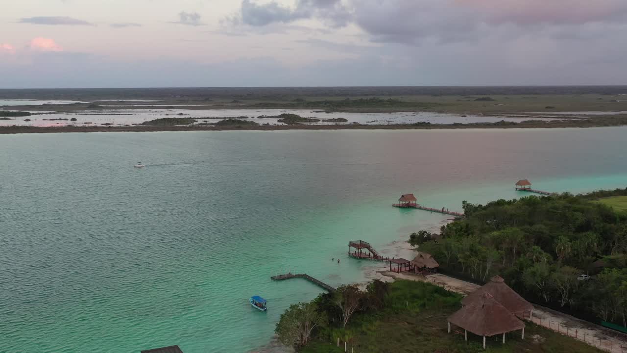 Smuk solnedgang over Bacalar-lagunen i Mexico med gyldent lys, der reflekteres på det stille vand. Silhuetten af kystlinjen og dramatiske skyer skaber et fredfyldt tropisk landskab i den gyldne time