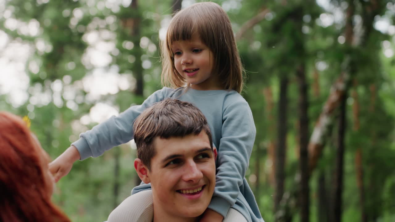 Un fratello porta la sorellina sulle spalle mentre la madre cerca scherzosamente di metterle un fiore tra i capelli; la bambina si volta timidamente con un sorriso, creando una scena toccante di tenerezza familiare all'aperto