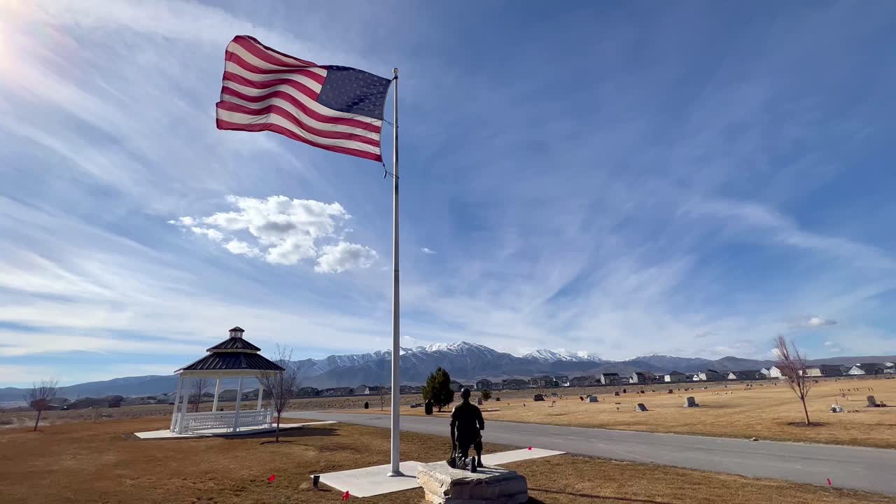 una bandera ondea en la brisa en un monumento conmemorativo de guerra ubicado en un cementerio con montañas en el fondo y un cielo rayado de nubes