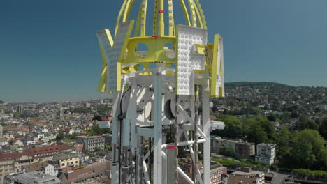 Toma-Aérea-De-Drones-Orbitando-Alrededor-De-La-Torre-De-Caída-Libre-Del-Parque-De-Diversiones-Y-Volando-Hacia-Atrás-Revelando-La-Ciudad-De-Zúrich,-Suiza-Durante-Zürichfest