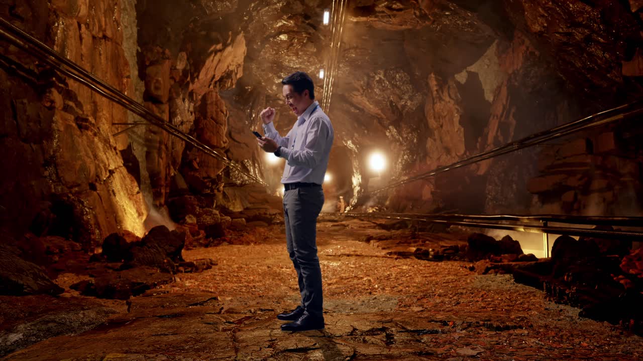 Full Body Side View Of An Asian Male Professional Worker Standing With His phone In Underground Mine Tunnel, He Raises His Fist Up With Screaming Goal