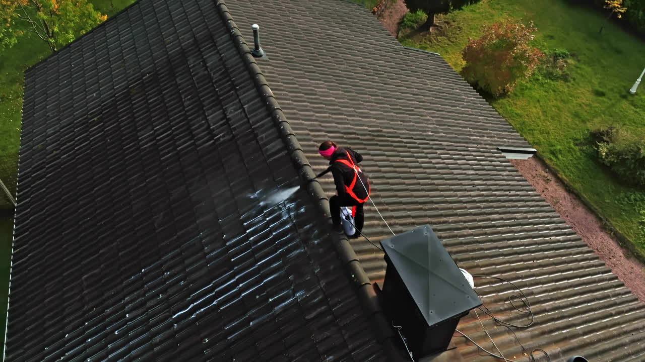 Drone rising in front of a woman pressure washing a house roof, sunny, fall day