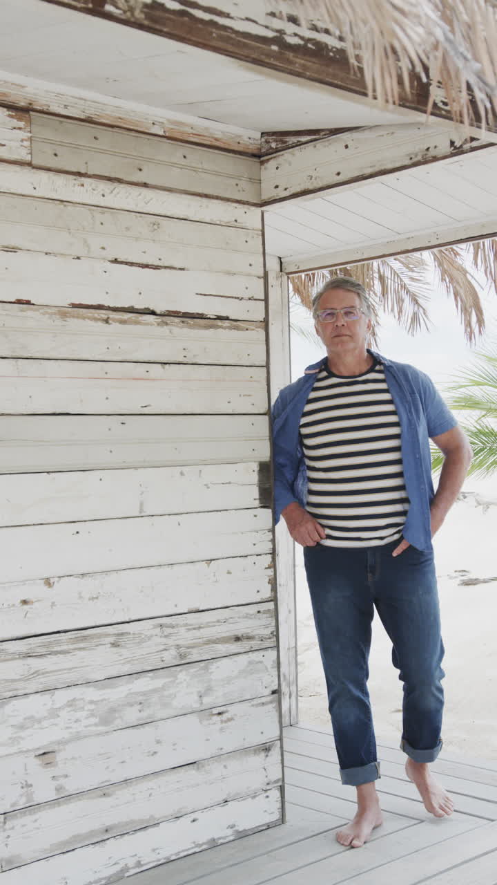 Vertical video of thoughtful senior caucasian man walking by beach hut on sunny beach