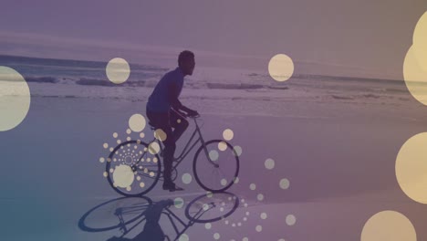 Yellow-spots-of-light-floating-against-african-american-man-riding-a-bicycle-at-the-beach