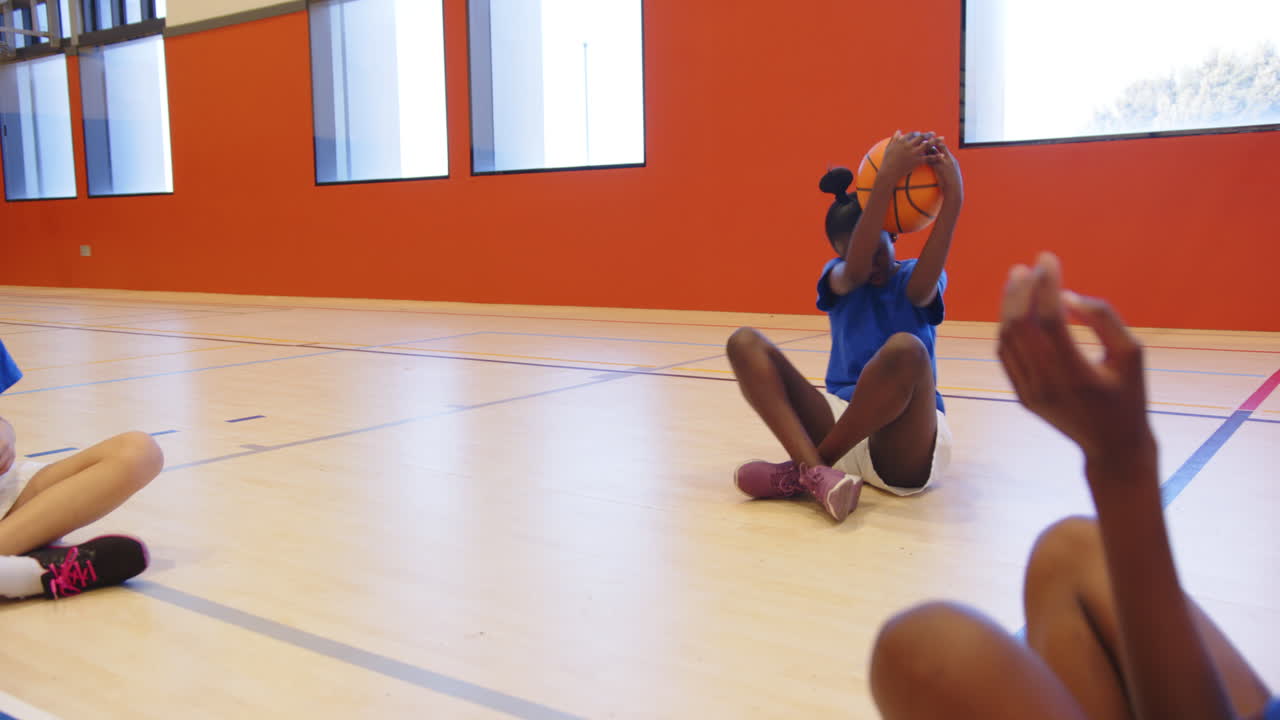Playing basketball in school gym, girls sitting on floor passing ball