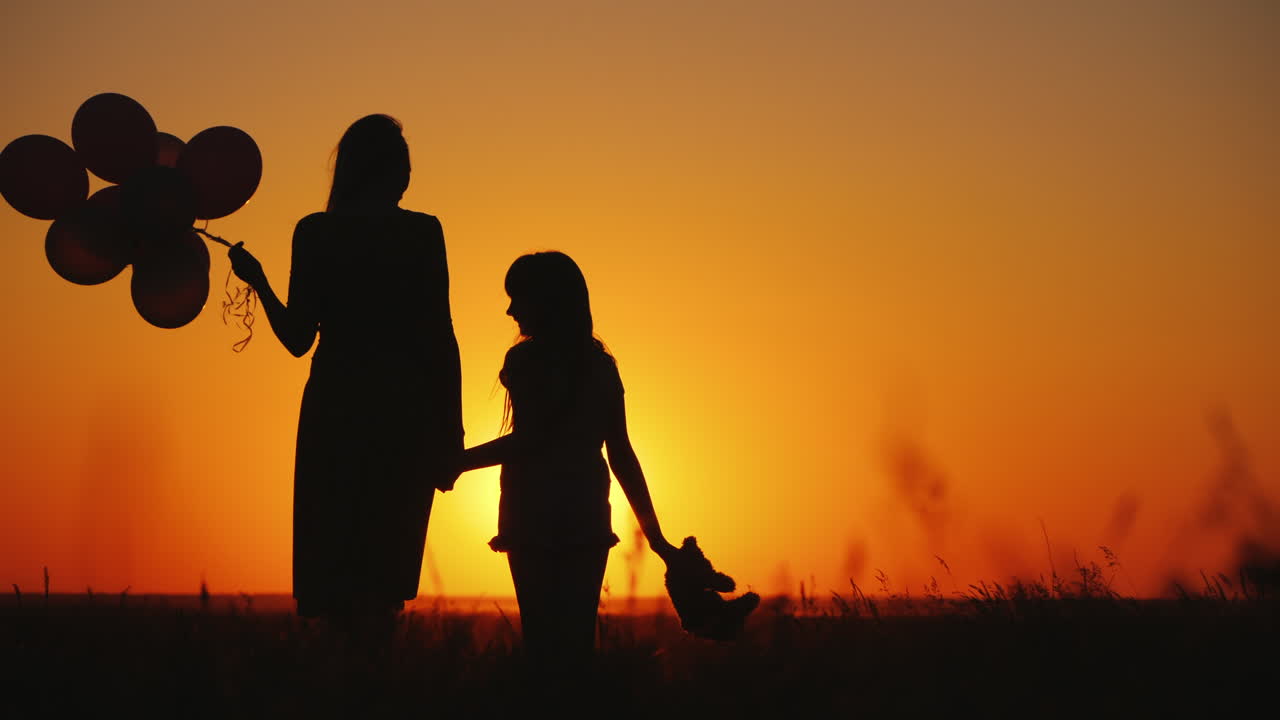 엄마와 딸은 일몰 때 초원에 서 있습니다. 그들은 공과 곰인형을 들고 있습니다.