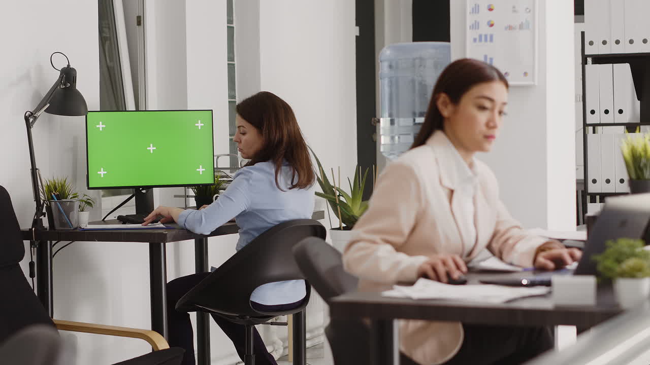 Two women working at their desks in an office with a computer and green screen