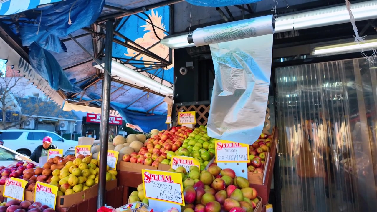 New York, USA, 20 dicembre 2025: Esposizione di mercato di frutta all'aperto con mele e manghi. Casse di prodotti colorati riempiono una bancarella sul marciapiede sotto tende da sole blu e cartelli con prezzi scritti a mano.