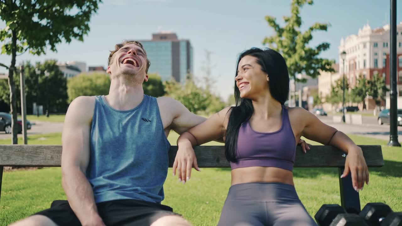 Uomo e donna che ridono in abbigliamento sportivo si scambiano gesti affettuosi su una panchina del parco dopo l'allenamento, con manubri.