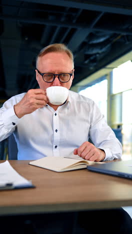 man takes a short coffee break from work in the office. portrait of a busy male sitting at desk drinking from cup. vertical video.