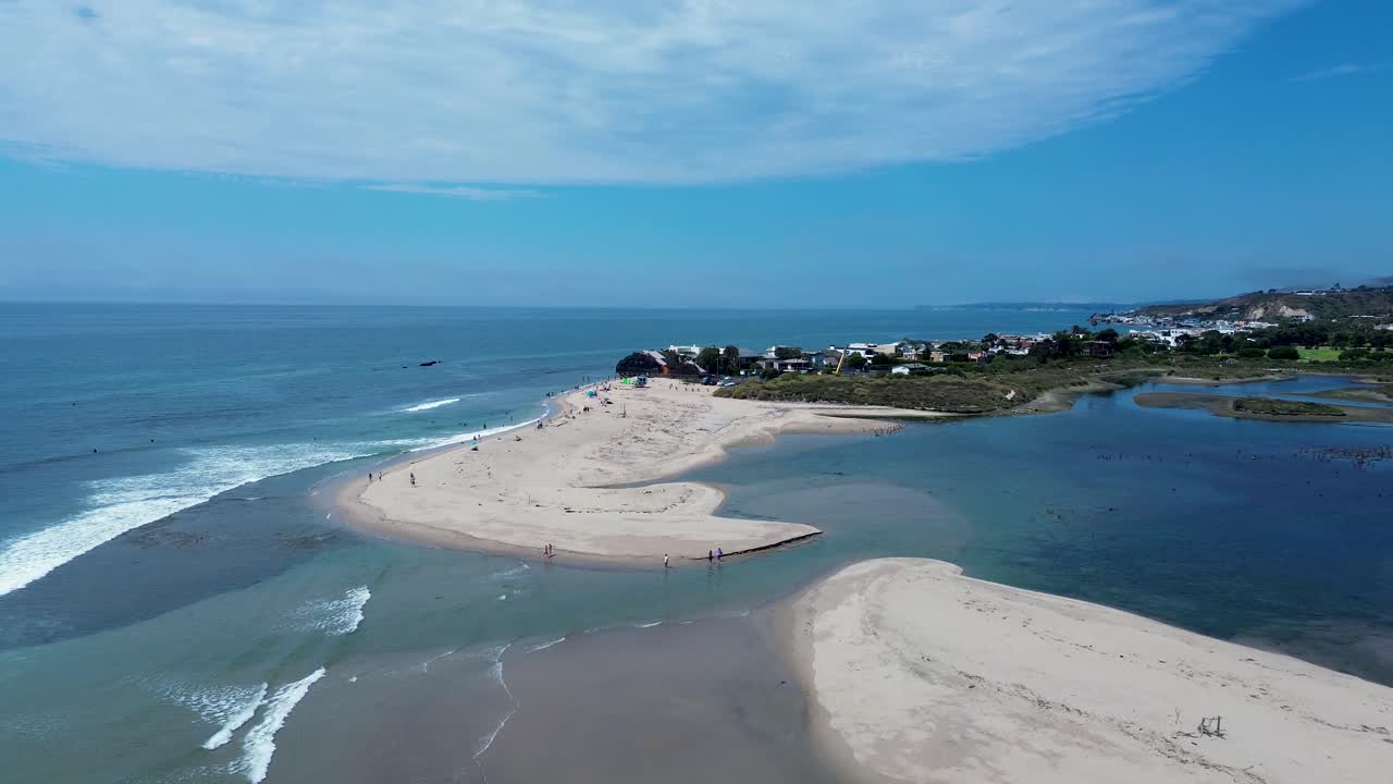 Aerial view of a beach, ocean, and lagoon where a river meets the sea, with a coastal town in the background