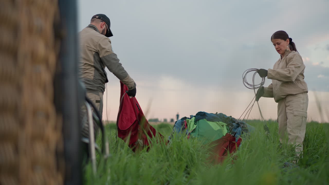 friends setting up for hot air balloon flight walking across grassy field dragging colorful envelope material and steel lines during preflight under cloudy sky in rural landscape