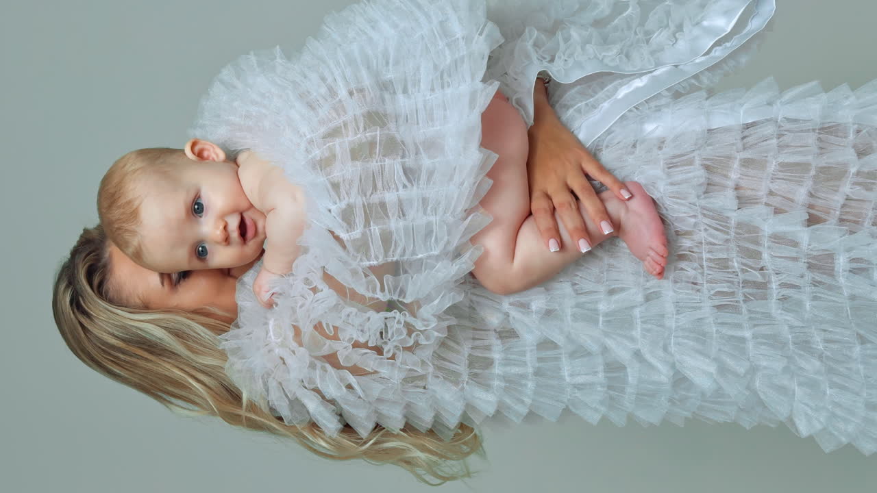 Lady in white robe presses her baby to the heart. Mom kisses her son yawning adorably. Vertical screen.