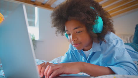 boy in bedroom lying on bed wearing wireless headphones using laptop computer